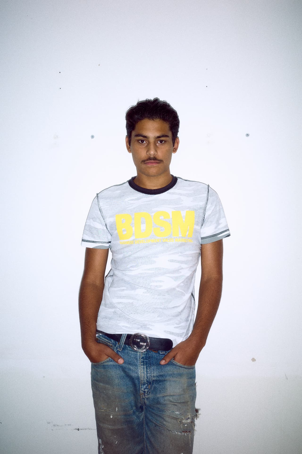 Model wearing a Connie Costas camouflage t-shirt with yellow BDSM lettering and denim jeans, photographed by Louis Flashman